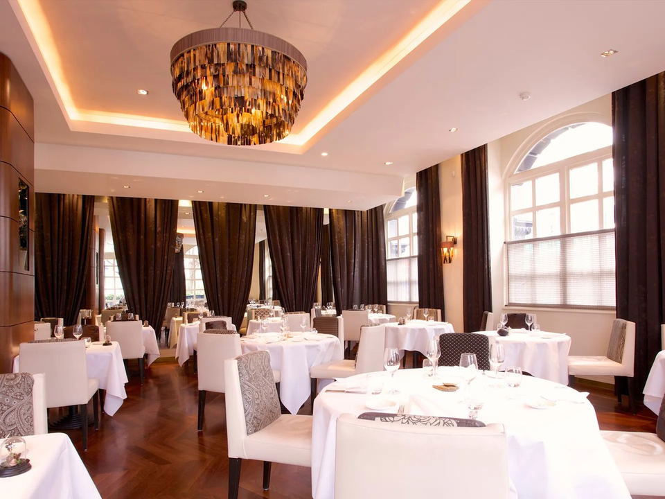 An elegant fine-dining room with white-clothed tables, white upholstered chairs, patterned cushions, a large tiered amber chandelier, dark curtains,