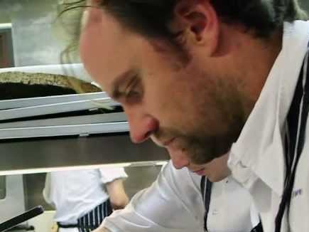 Chef, wearing a white jacket and striped apron, plates a dish on a metal tray in a bustling professional kitchen.