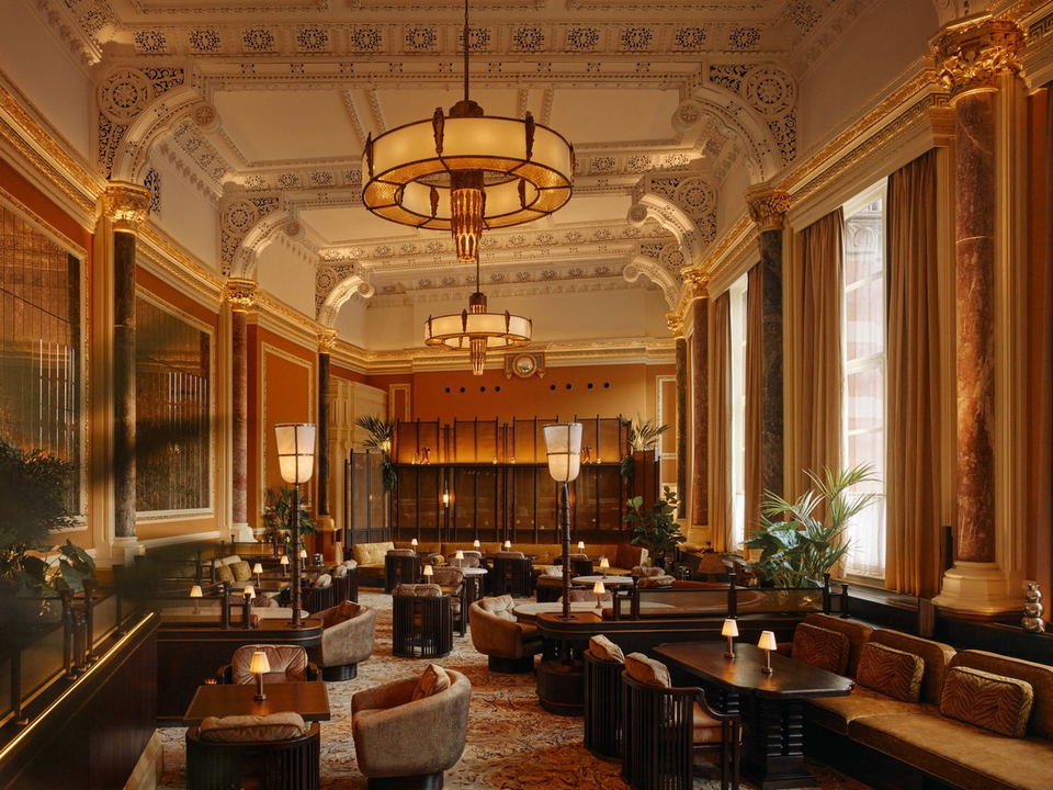 A grand dining room with ornate white plasterwork ceilings, gilded columns, amber walls, large pendant chandeliers, and plush seating.