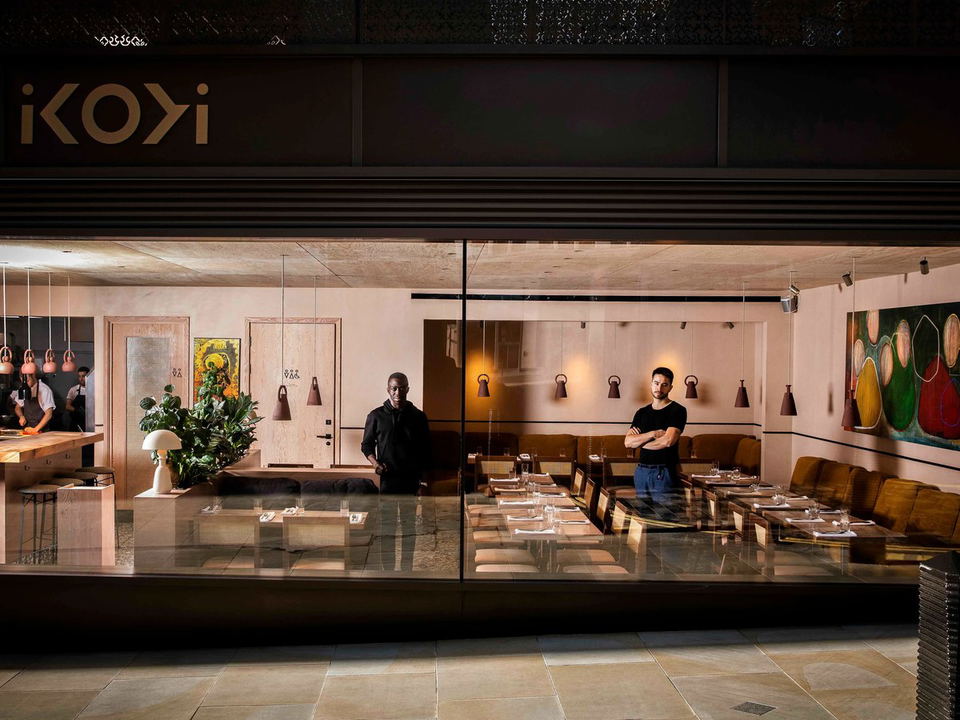 The interior of Ikoyi restaurant at 180 Strand, with its 'ikoyi' signage, two men, and dining tables visible through a glass facade.