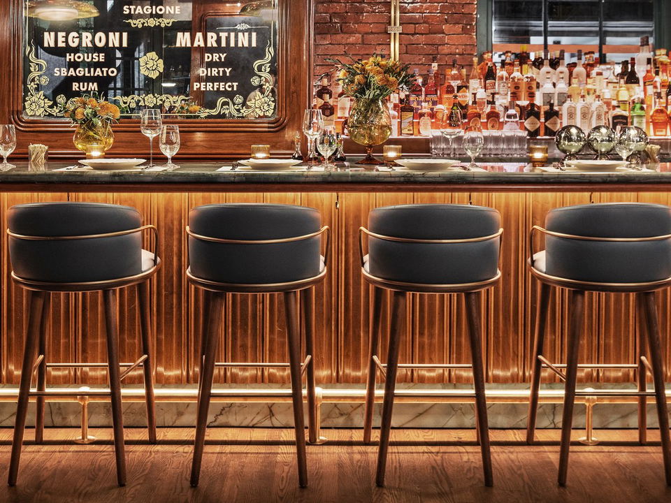 An elegant bar interior features a row of navy-and-brass bar stools, a marble base, a dark stone counter, and a backbar with spirits and a