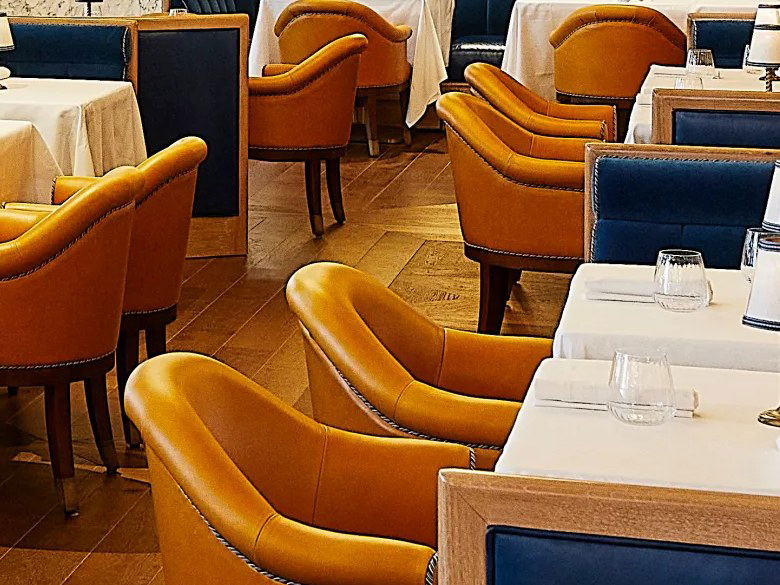 An elegant restaurant dining room interior featuring large indoor trees, tiered brass chandeliers with globe bulbs, deep blue velvet banquette