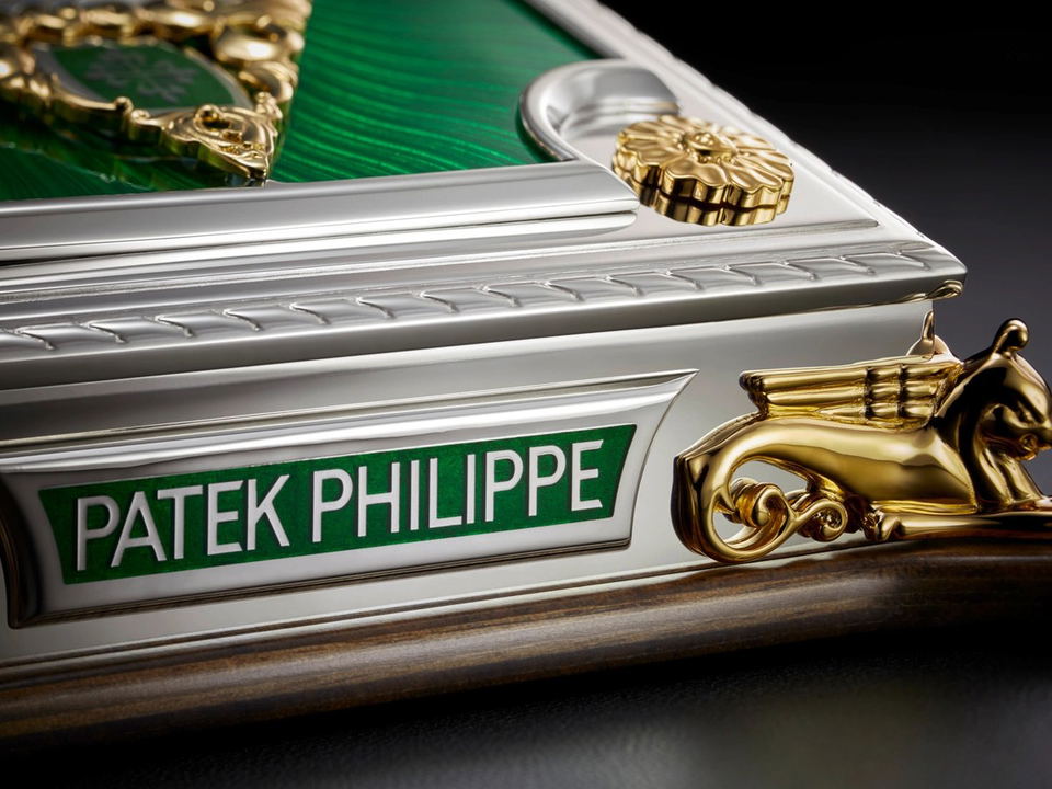 A close-up of a Patek Philippe display case featuring a green enamel surface, silver metalwork, and gold griffin figurines.