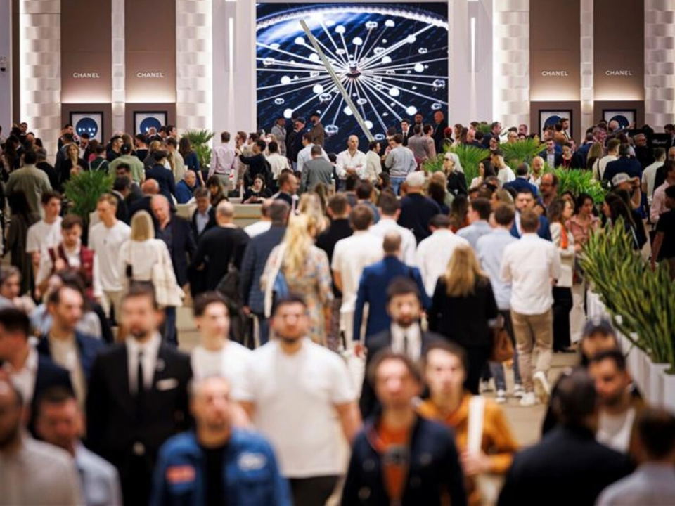 Crowd of attendees at a watch exhibition with Chanel displays in the background.