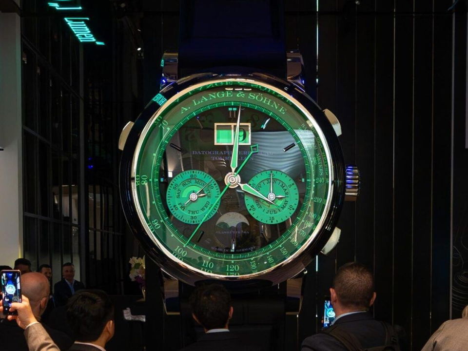An oversized A. Lange & Söhne Datograph watch with a luminous green dial and strap is displayed, surrounded by attendees at Watches and Wonders.