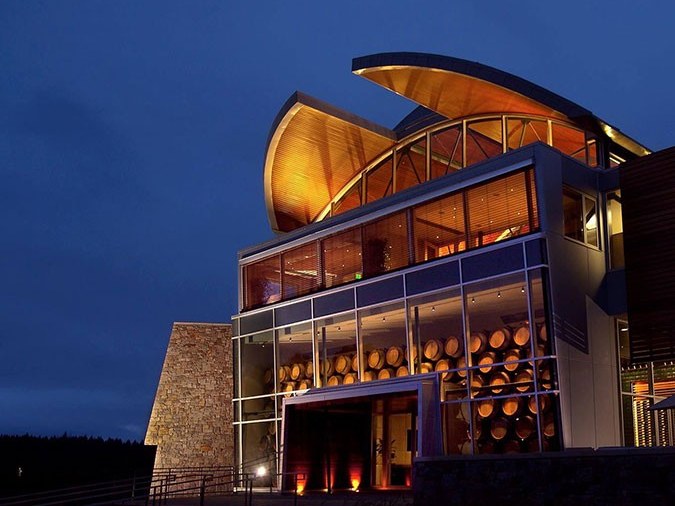 A modern winery building at night with a curved, illuminated wood-paneled roof, glass facade, and visible wine barrels inside.