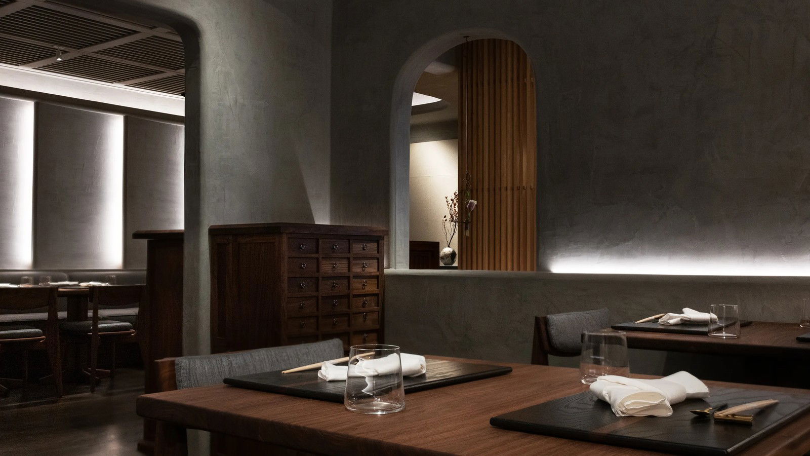 An elegant, dimly lit Japanese restaurant dining room with dark gray shikkui lime plaster walls, walnut furniture, and arched doorways.