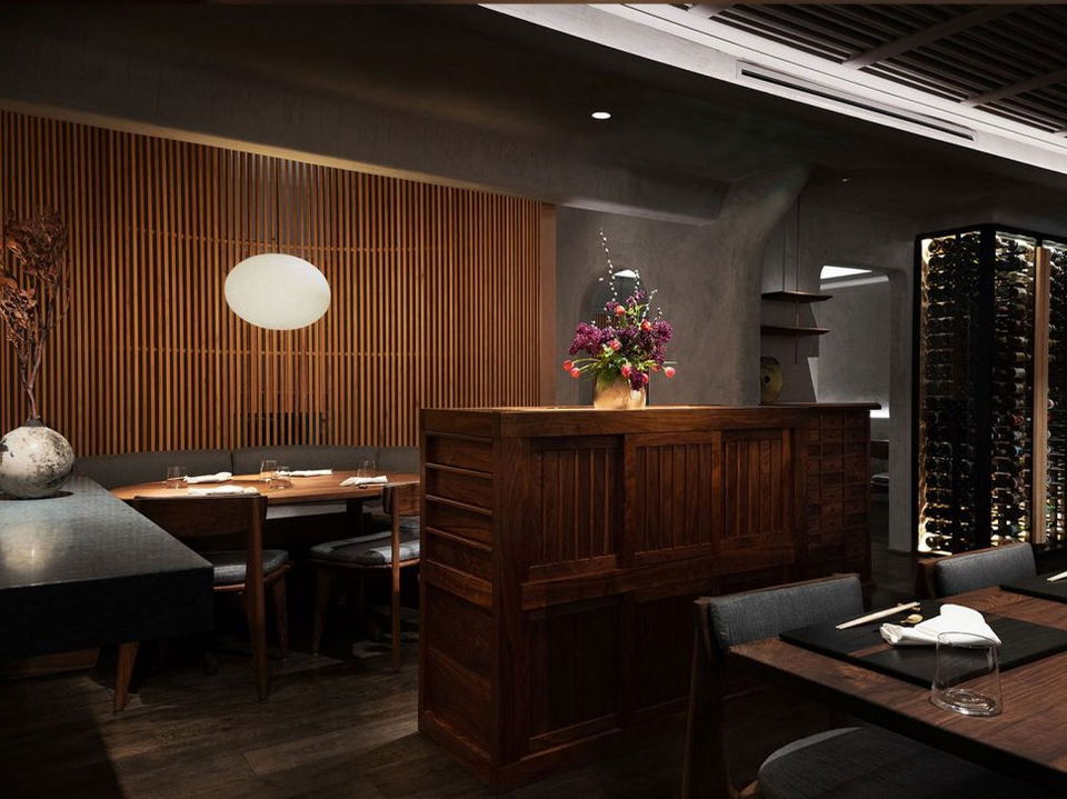 An elegant restaurant interior features concrete walls, warm wood panels, a dark wooden host stand with flowers, and a glass wine cellar.