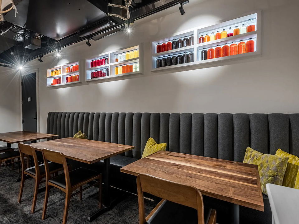 A modern restaurant dining room with a long gray banquette, wooden tables, and illuminated wall niches displaying colorful bottled beverages.