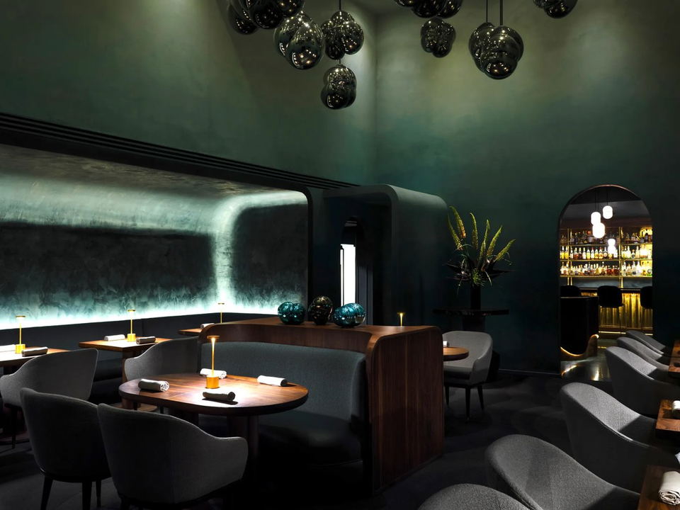 An elegant, dimly lit dining room with deep teal walls, curved banquettes, dark wood tables with small lamps, and mirrored pendant lights.