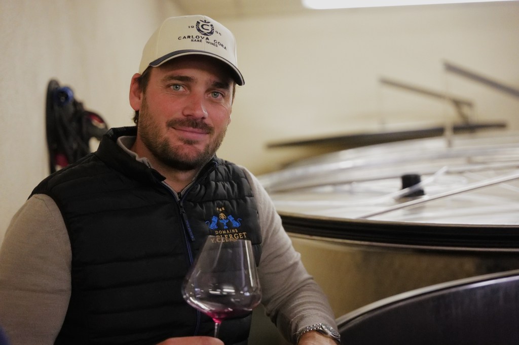 A man in a black vest holding a wine glass in a cellar with barrels visible in the background.