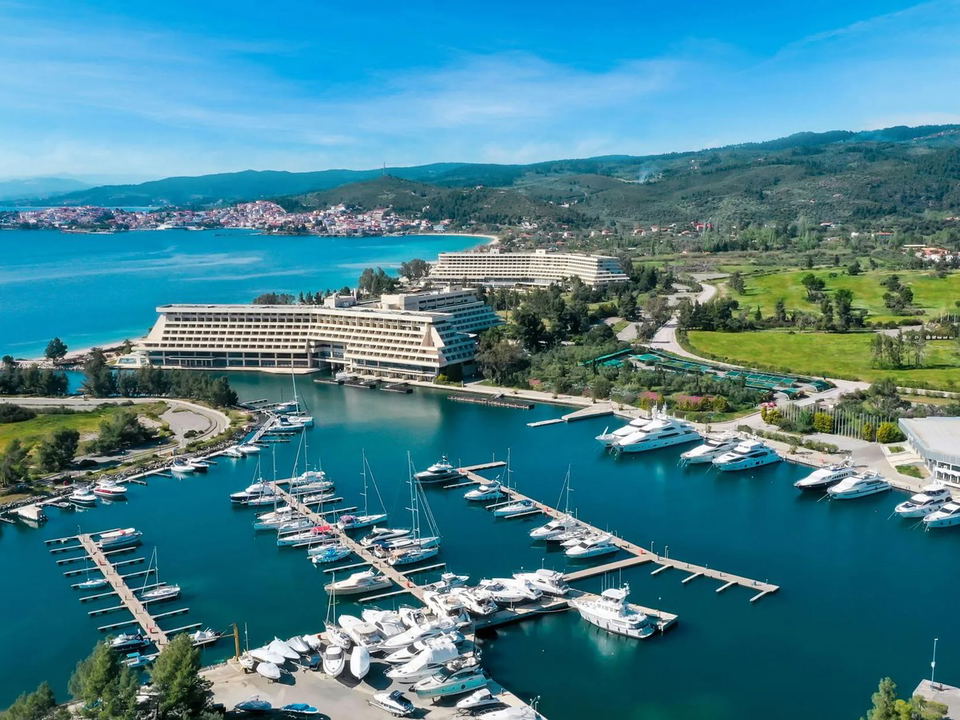 An aerial drone view of Porto Carras Grand Resort's marina, filled with numerous yachts and sailboats, with terraced hotel buildings behind.