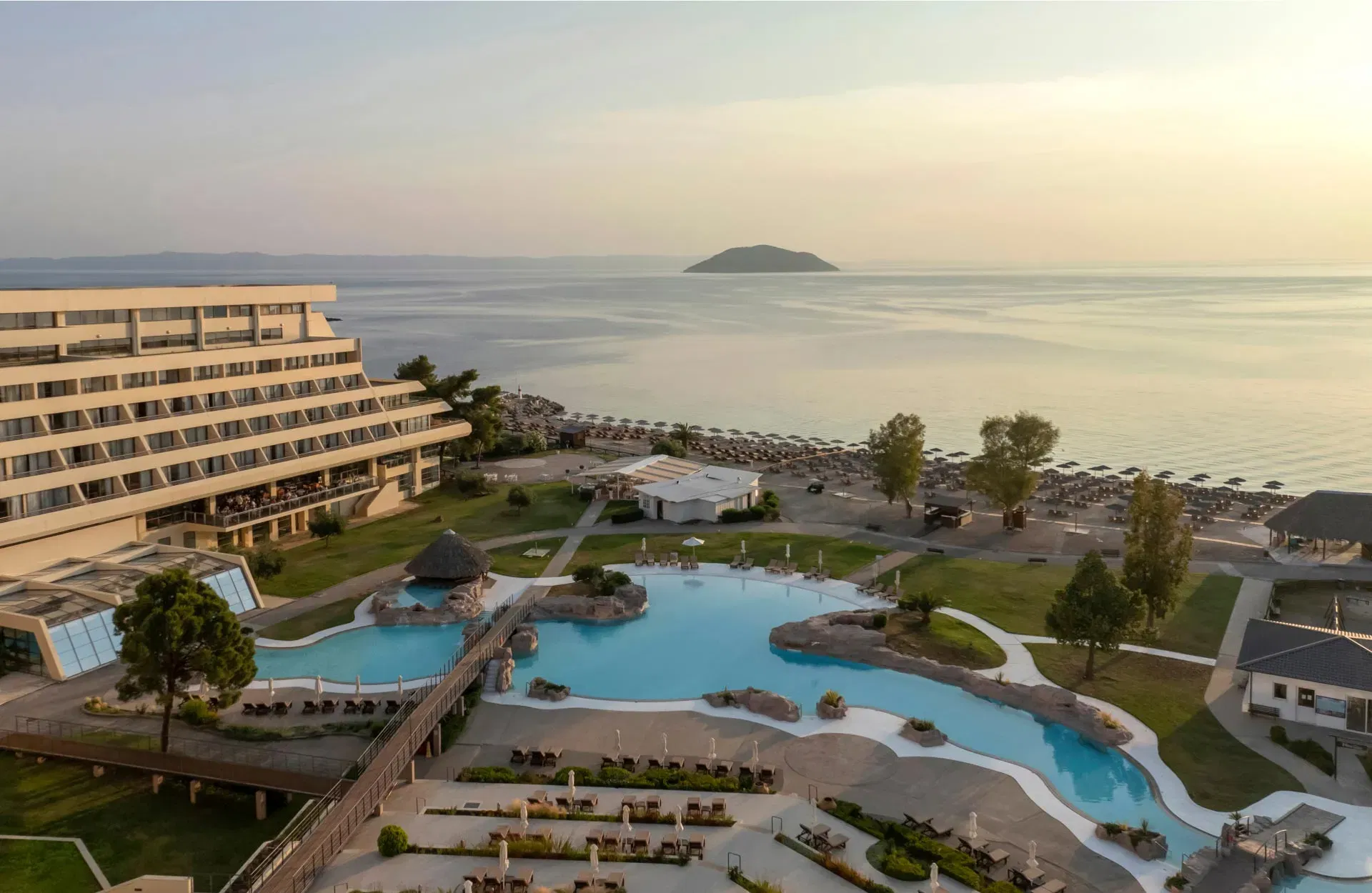 An aerial view of the Porto Carras Meliton Hotel, featuring a large, multi-level building with balconies, a complex of swimming pools with a bridge,