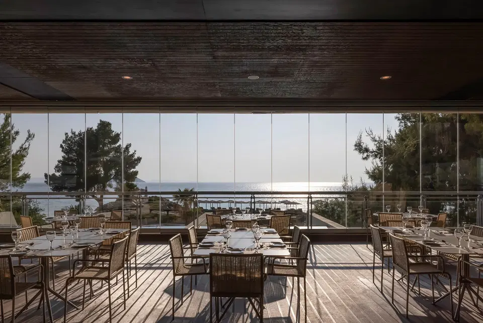 An empty restaurant dining room with tables set for guests, overlooking a calm sea and distant land under a bright sky.