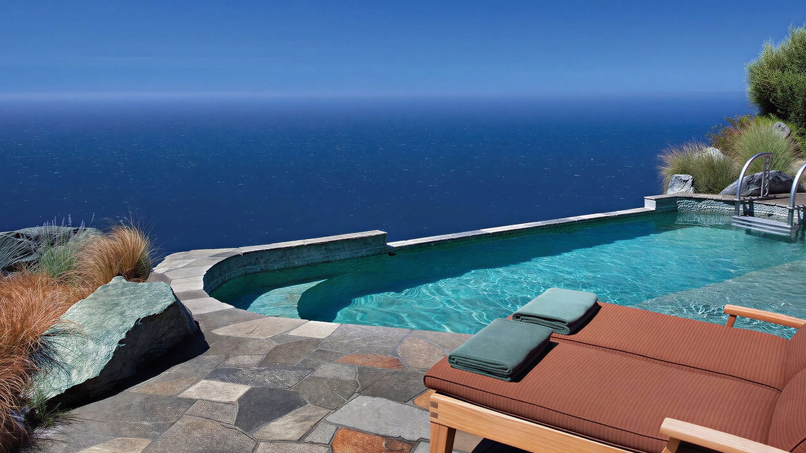An infinity pool with turquoise water overlooking a vast blue ocean under a clear sky, with a lounge chair and towels.