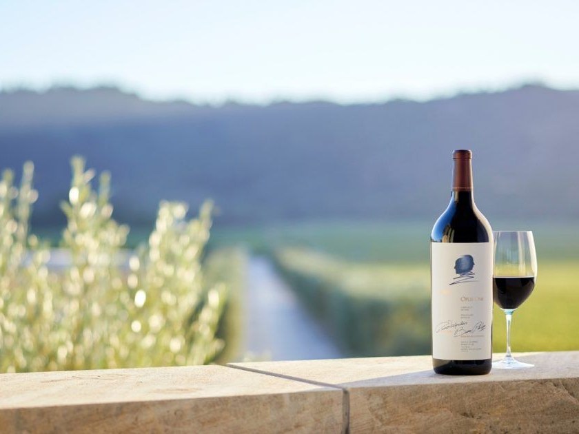A bottle of Opus One wine and a filled wine glass sit on a stone ledge overlooking vineyards and mountains.