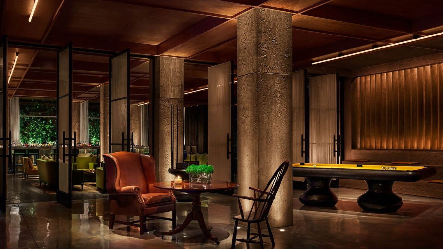 A dimly lit, luxurious lounge area with a pool table, comfortable seating, and a bar area visible through glass partitions.