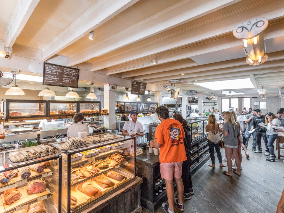 Crowded deli counter at Gjusta in Venice, California, with glass cases displaying meats, fish and pastries and overhead sandwich menu boards