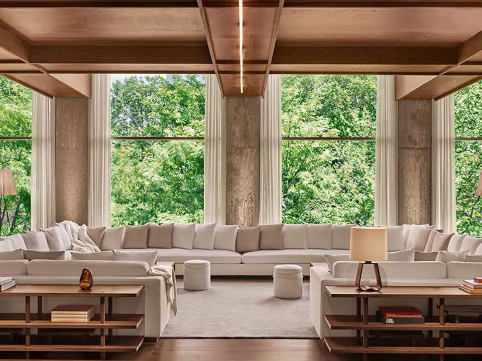 A spacious living room with a large U-shaped sofa, two end tables with books and lamps, and tall windows overlooking lush green trees.