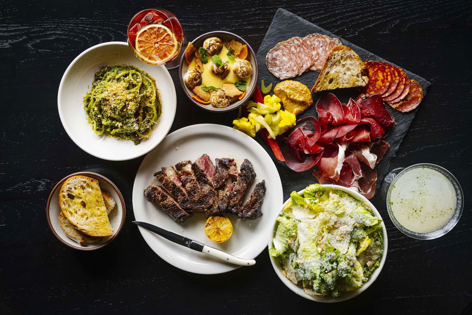 Overhead spread of Gjelina dishes including sliced grilled steak, charcuterie board, green herb pasta, Caesar salad, and a cocktail on a dark wood table.