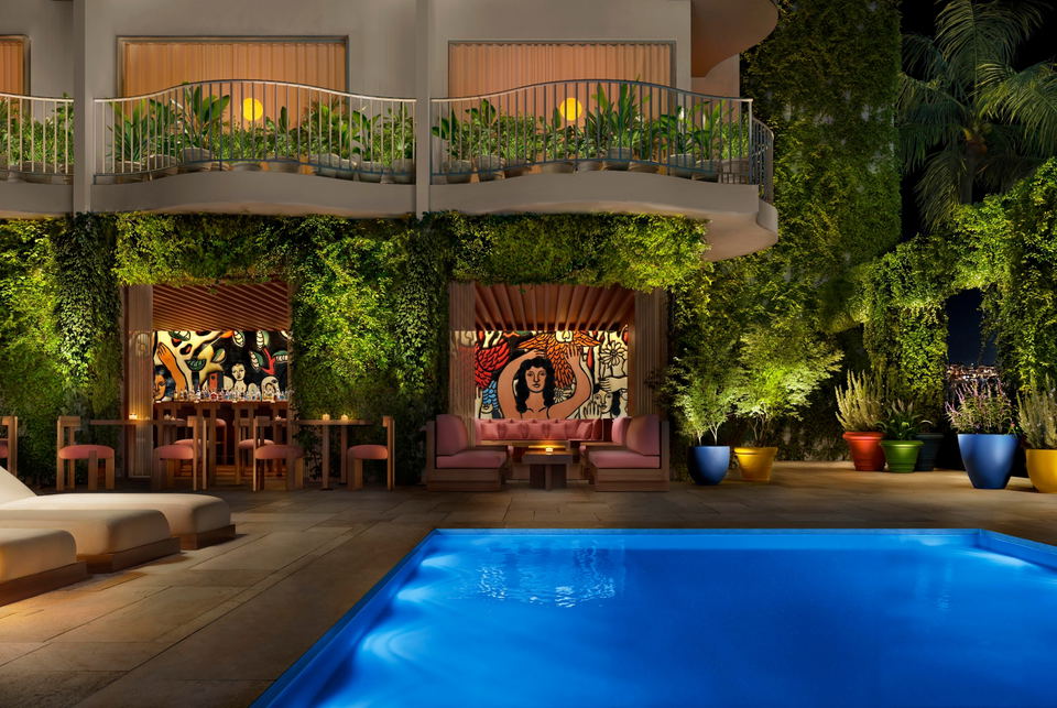 Illuminated outdoor pool at Public West Hollywood hotel, surrounded by a lush living green wall, colorful murals, pink lounge seating, and a bar at night.