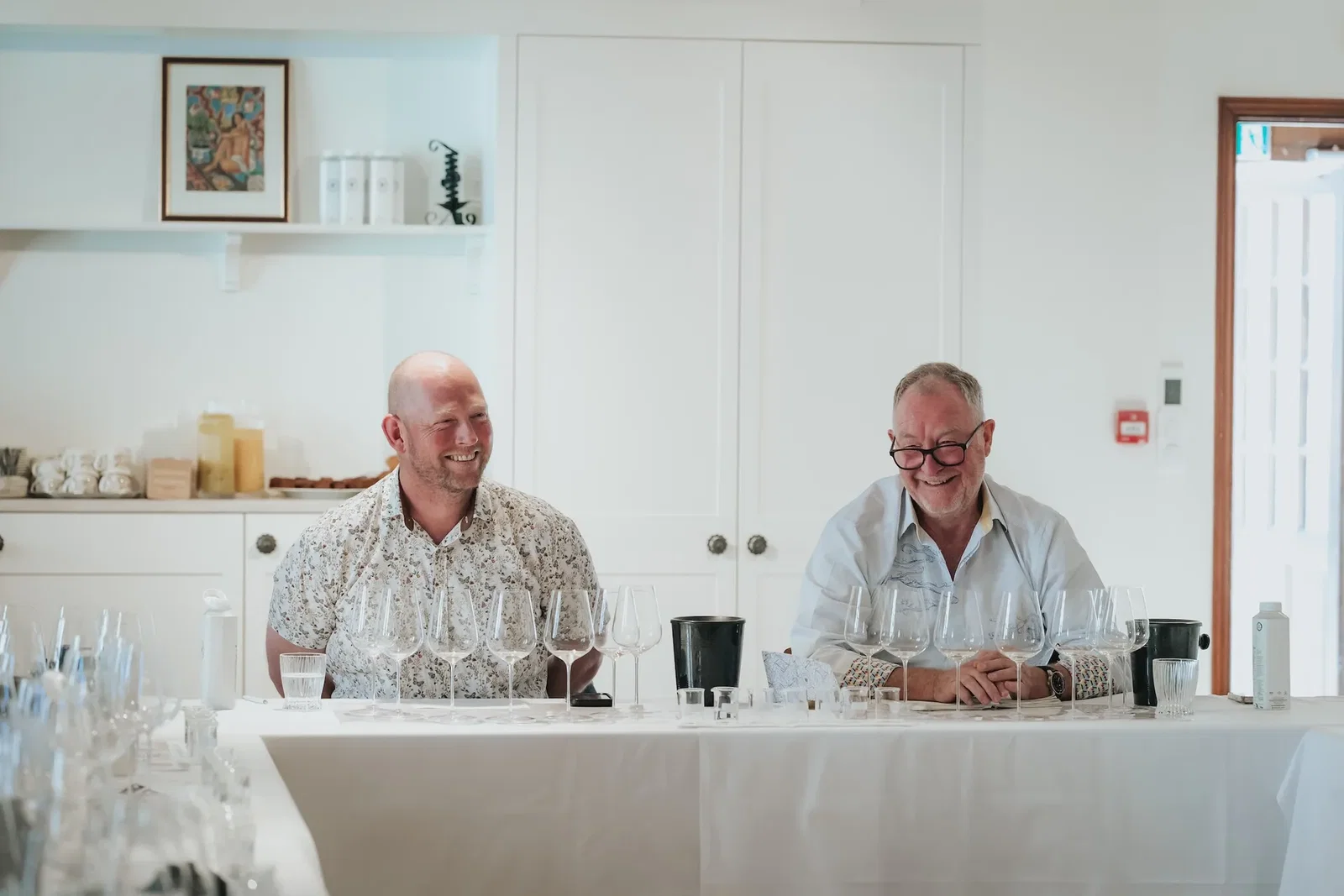 Two smiling men seated at a white table with multiple wine glasses in front of them in a modern white interior space.