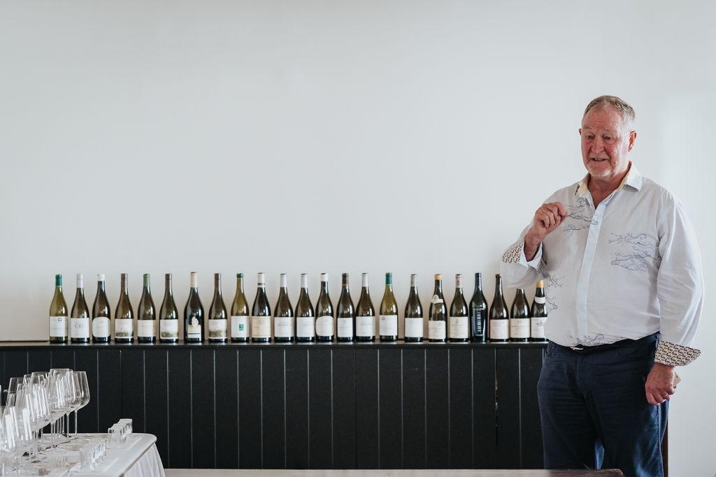 An older man in a light-colored shirt stands next to a long row of white wine bottles displayed on a black shelf against a white wall, with wine glasses visible in the foreground.