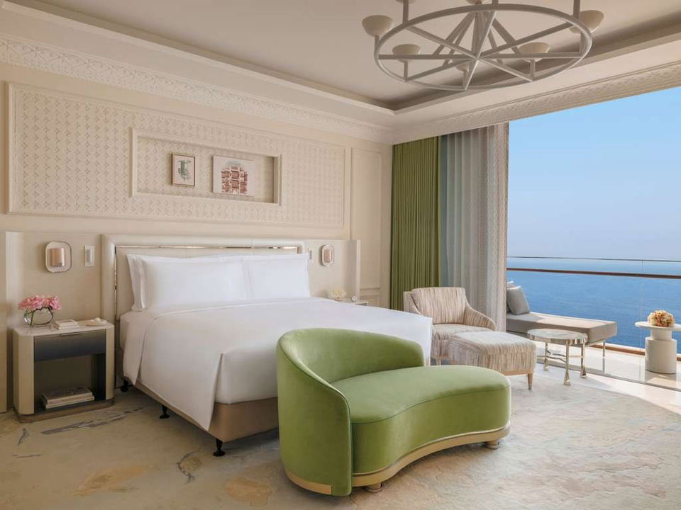 A luxurious bedroom at Raffles Jeddah with a large bed, a green curved sofa, and a balcony overlooking the ocean.