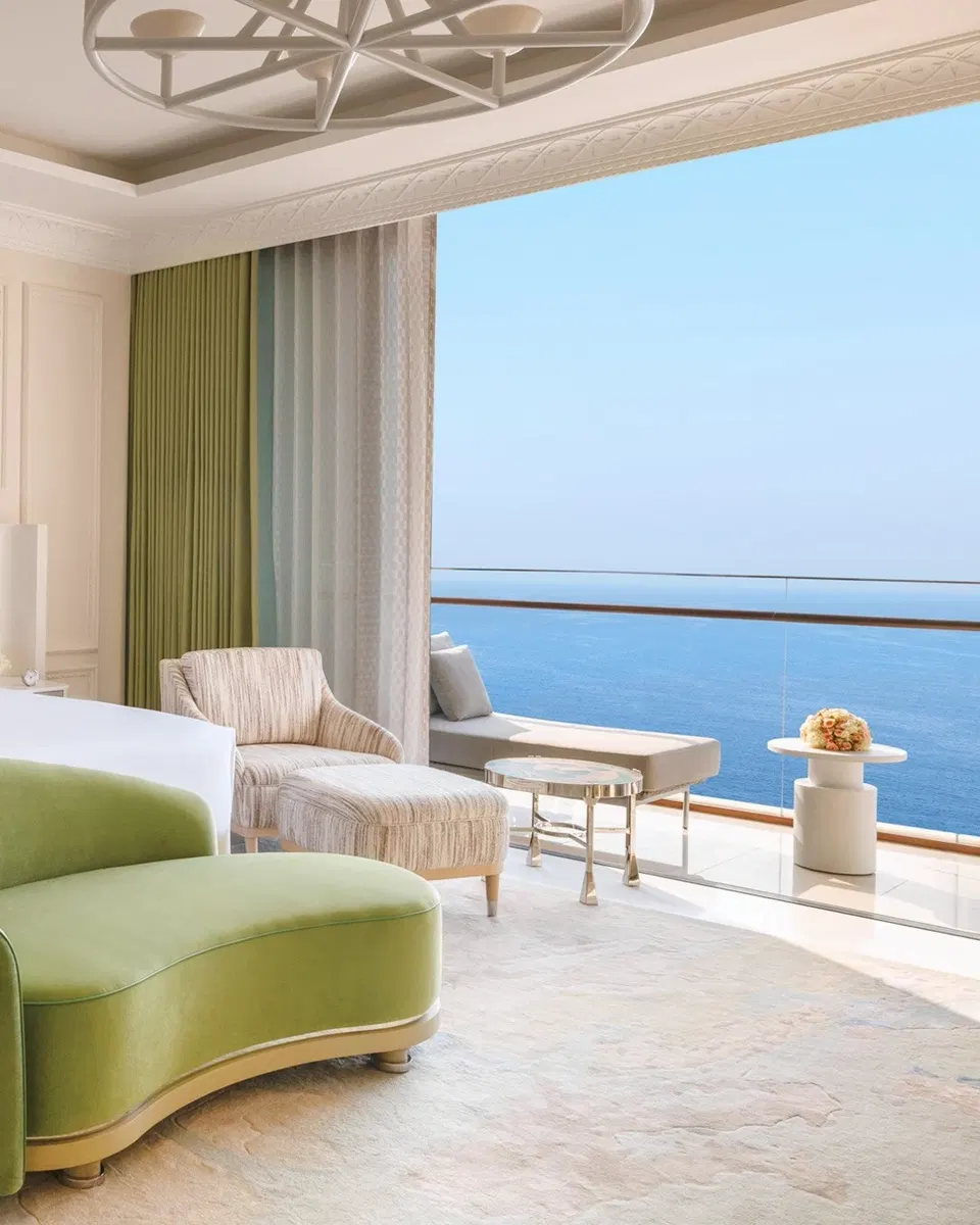 Luxury suite interior at Raffles Jeddah with a green curved sofa, woven armchair, and open balcony overlooking the Red Sea Corniche under a clear blue sky.