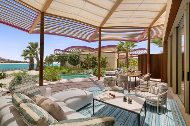 Modern resort terrace with comfortable seating, dining furniture, a pool, and retractable striped shade structures, with palm trees and water views in the background.