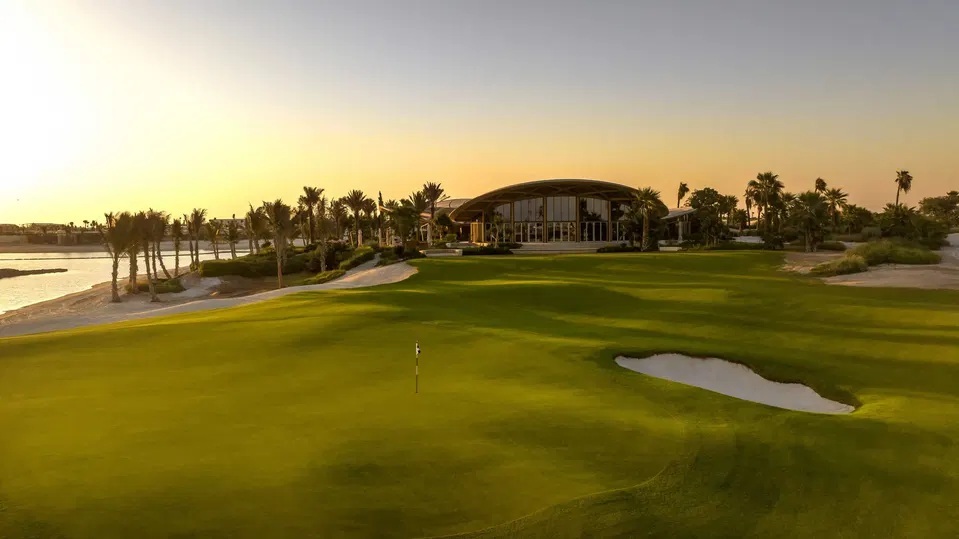 Golf course green with flag, sand bunker, palm trees, and modern curved clubhouse building at sunset beside water