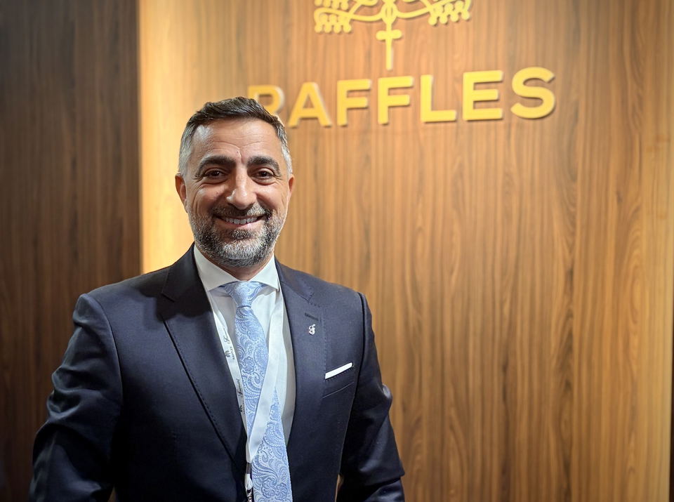Professional portrait of a man in a dark suit and light blue tie standing in front of a wooden wall with 'RAFFLES' signage
