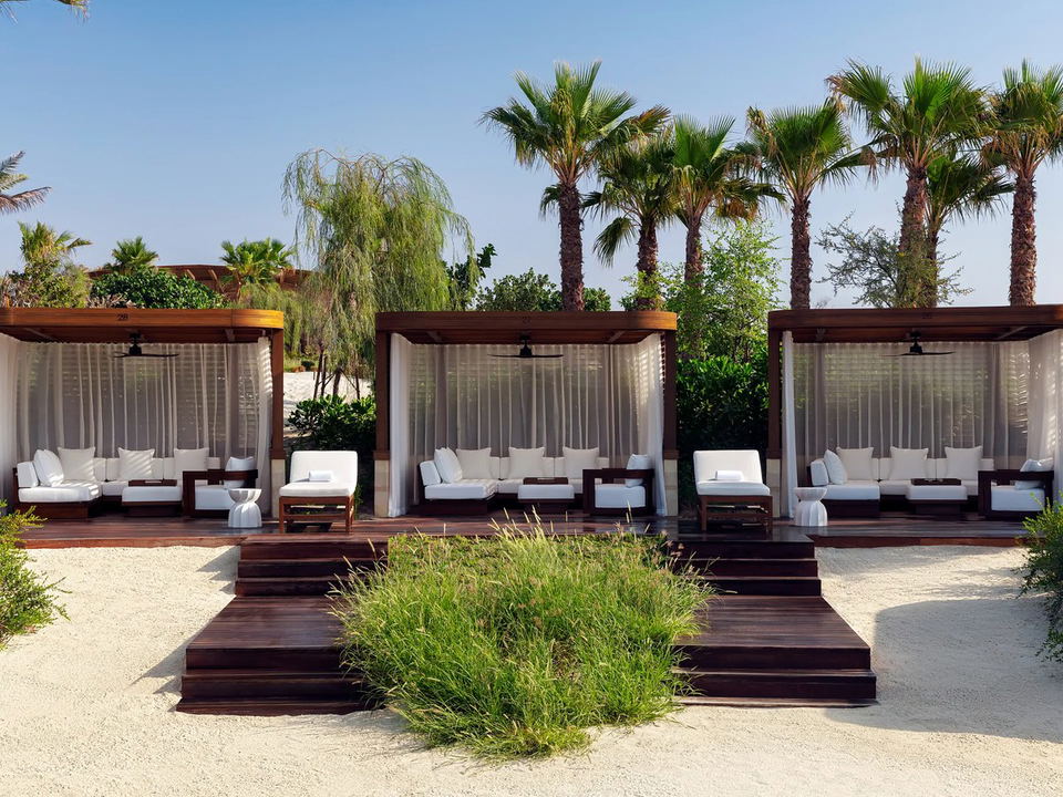Three luxury cabana structures with white curtains, dark wood frames, white daybeds, and palm trees in the background; published on Condé Nast Travele.