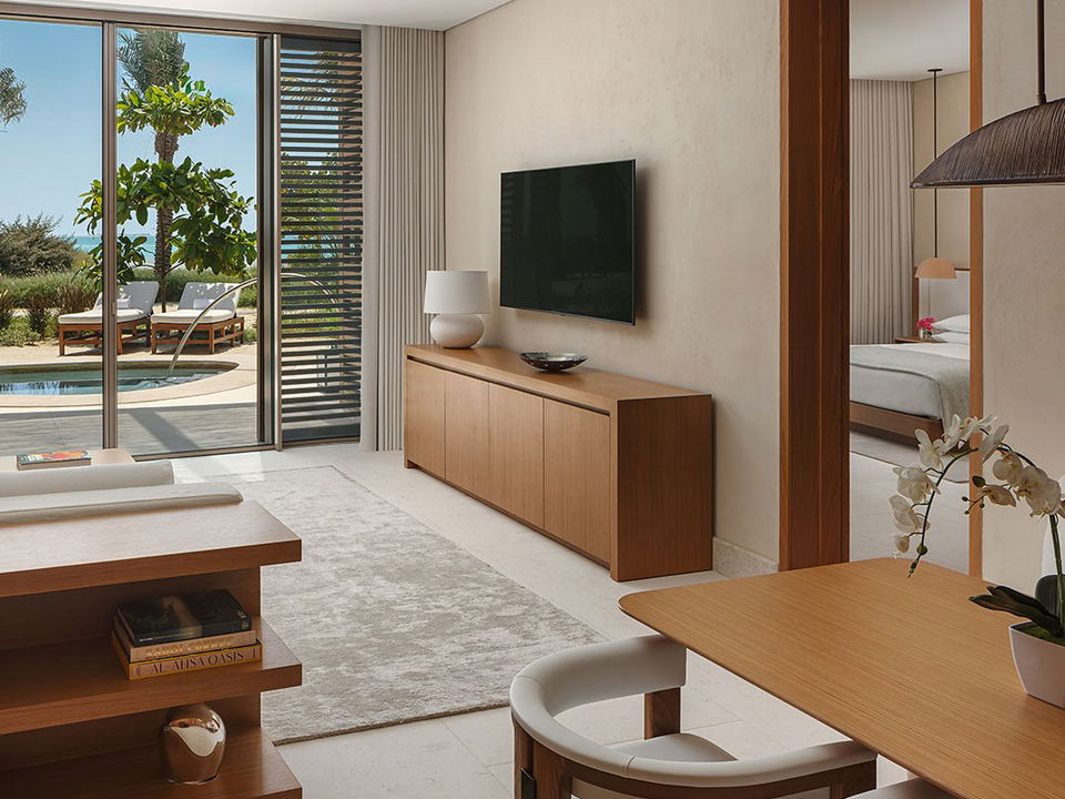 A modern hotel suite interior with natural oak furniture, coral stone flooring, and floor-to-ceiling windows opening to a pool terrace; the source.