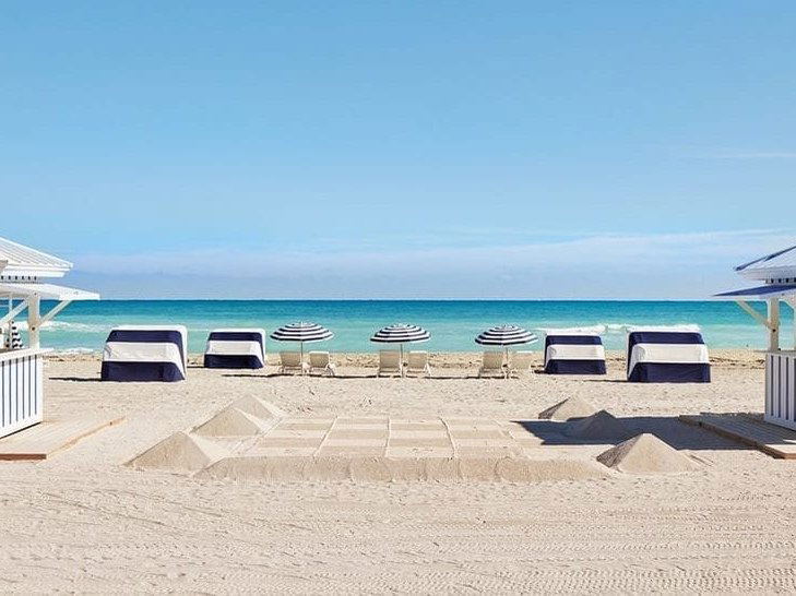 Beach scene with navy and white striped beach loungers, matching umbrellas, and white architectural structures flanking a sandy beach with clear turquoise water.