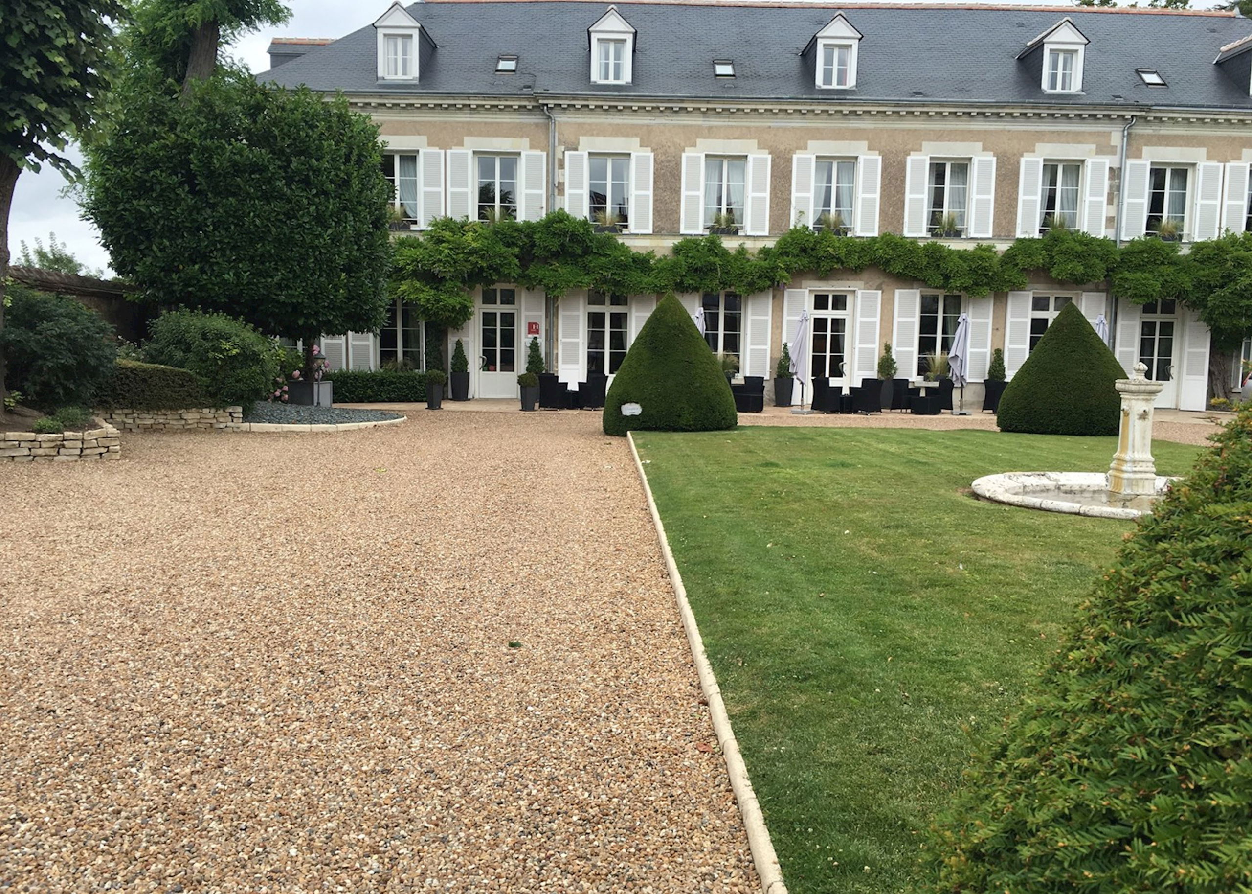 Elegant French manor house exterior with beige stone facade, white shutters, mansard roof with dormers, manicured lawn, topiary, and gravel courtyard;