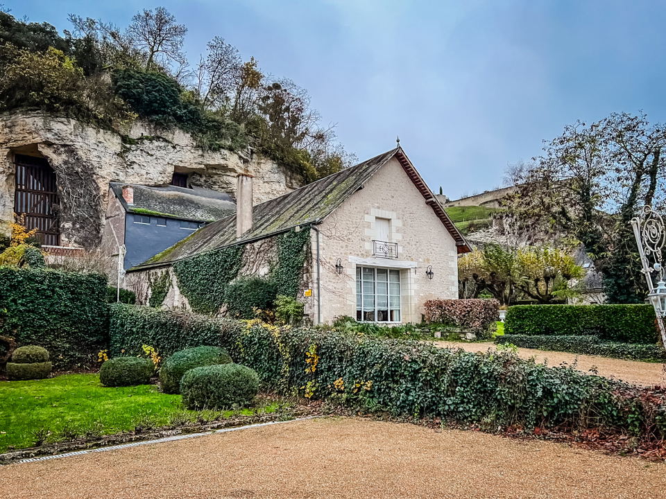 Exterior view of a stone building with gabled roof and landscaped garden, set against a hillside with cave dwellings; the source page is a review of H.