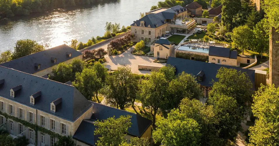 Aerial photograph of a multi-building estate complex with stone architecture situated on the banks of a river, surrounded by mature trees and landscaped grounds.