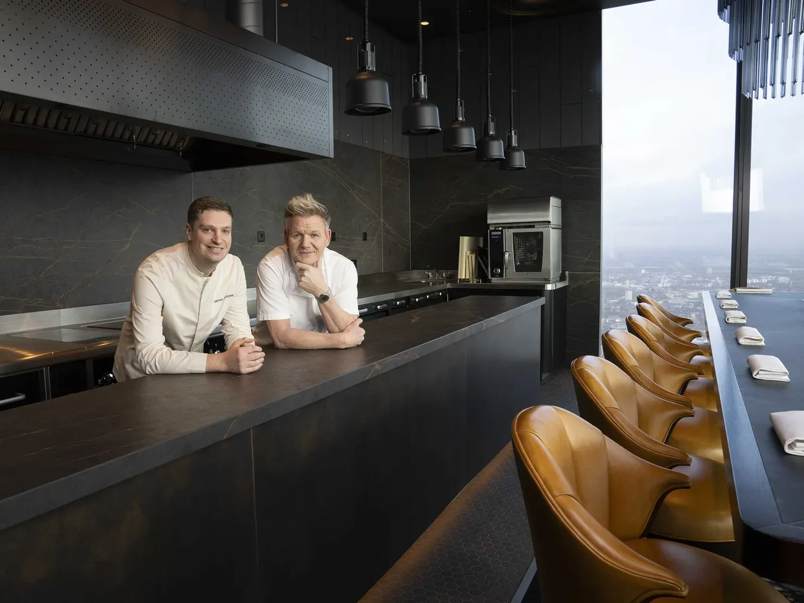 Two chefs, one with light brown hair and one with blonde hair, stand behind a dark counter in a modern kitchen with a city view.