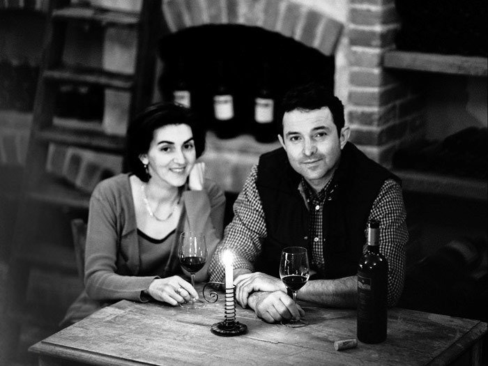 Black and white portrait of a couple seated at a wooden table with wine glasses and a bottle in what appears to be a wine cellar with brick archway; t
