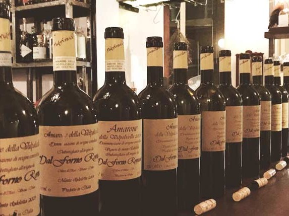 A row of dark glass wine bottles with light-colored labels, featuring "Amarone della Valpolicella" and "Dal Forno Romano" text.