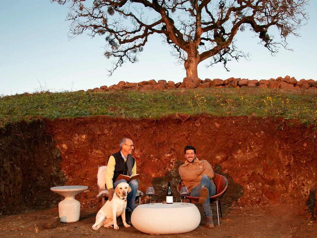 Two men sit in chairs inside a soil pit with red earth walls, a yellow Labrador beside them, a wine bottle and glasses on a low table, and a stone wall with a bare oak tree above.