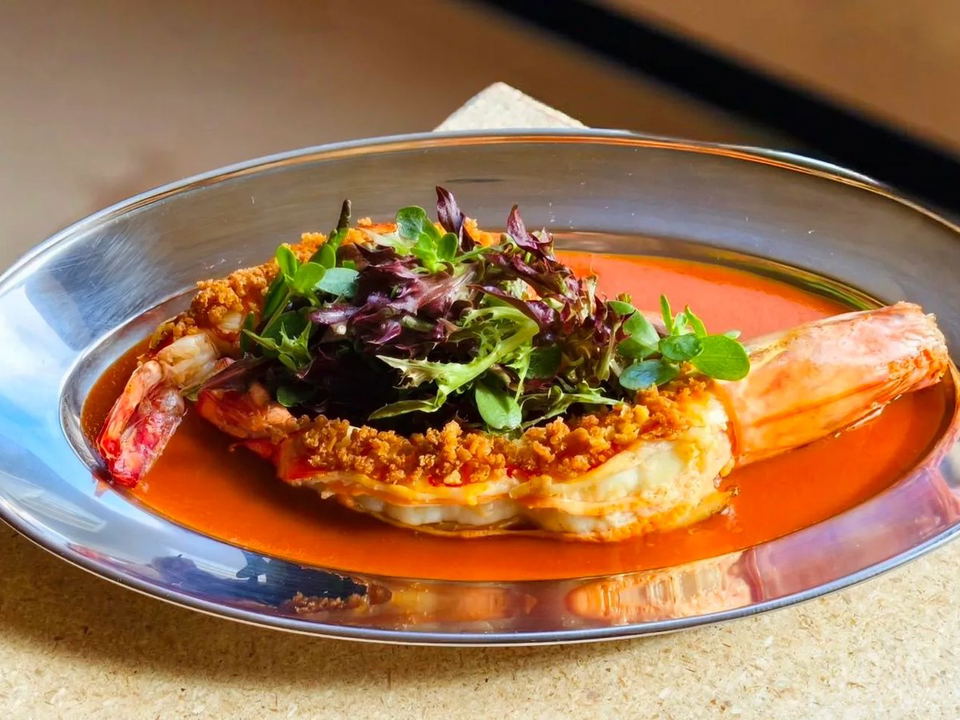 A vibrant shrimp dish, garnished with greens and crispy bits, rests in a rich orange sauce on a gleaming silver platter.