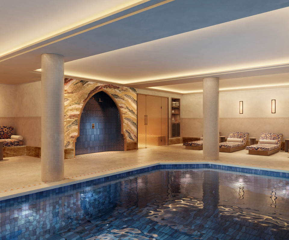 An indoor spa pool with blue tiled floor and walls, flanked by support columns, with a decorative arched doorway and cushioned lounge chairs visible in the background.