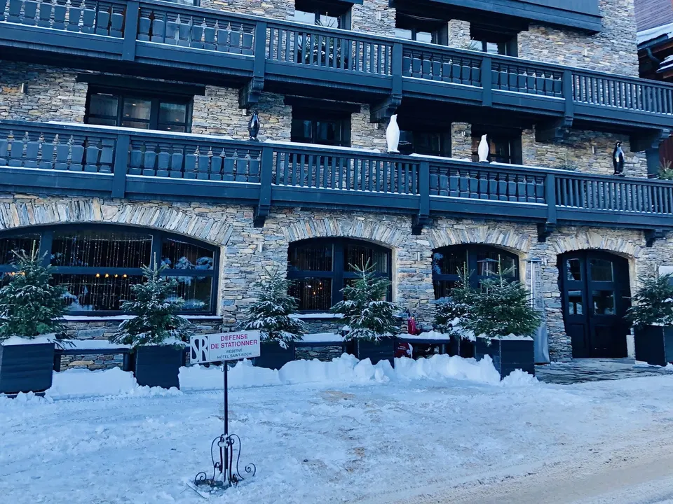 A multi-story stone building with blue wooden railings and balconies, featuring arched windows on the ground floor and snow on the ground in front.