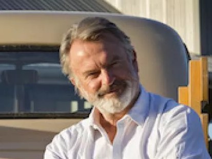 A middle-aged man with grey hair and facial hair wearing a light blue collared shirt, photographed outdoors with a vehicle visible in the background.