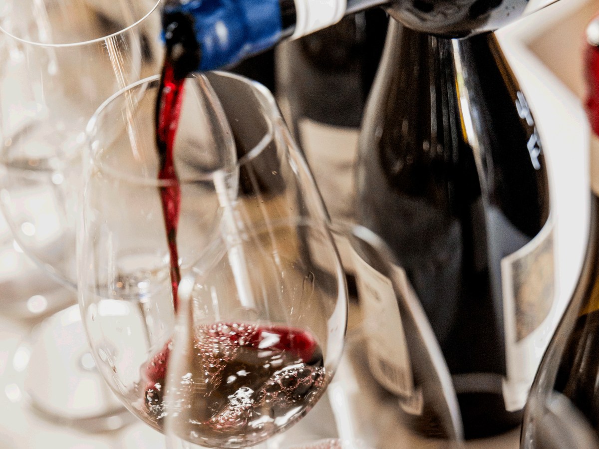 Red wine being poured from a bottle with a blue foil top into a clear glass, with other wine bottles in the background.