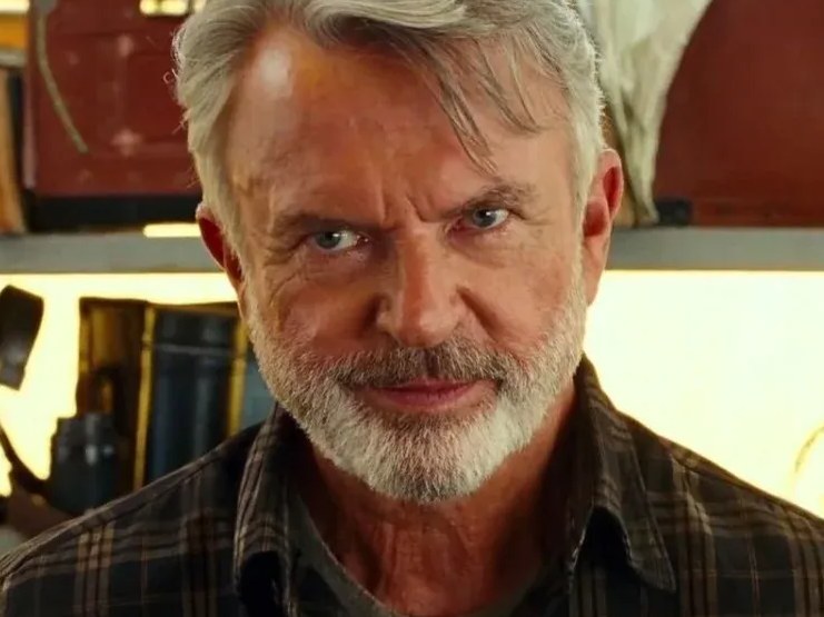 A close-up portrait of an older man with gray hair and a beard, wearing a plaid shirt, looking intently at the camera.