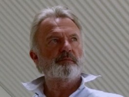 Portrait of an older man with a grey beard wearing a white shirt, photographed indoors against a plain wall.