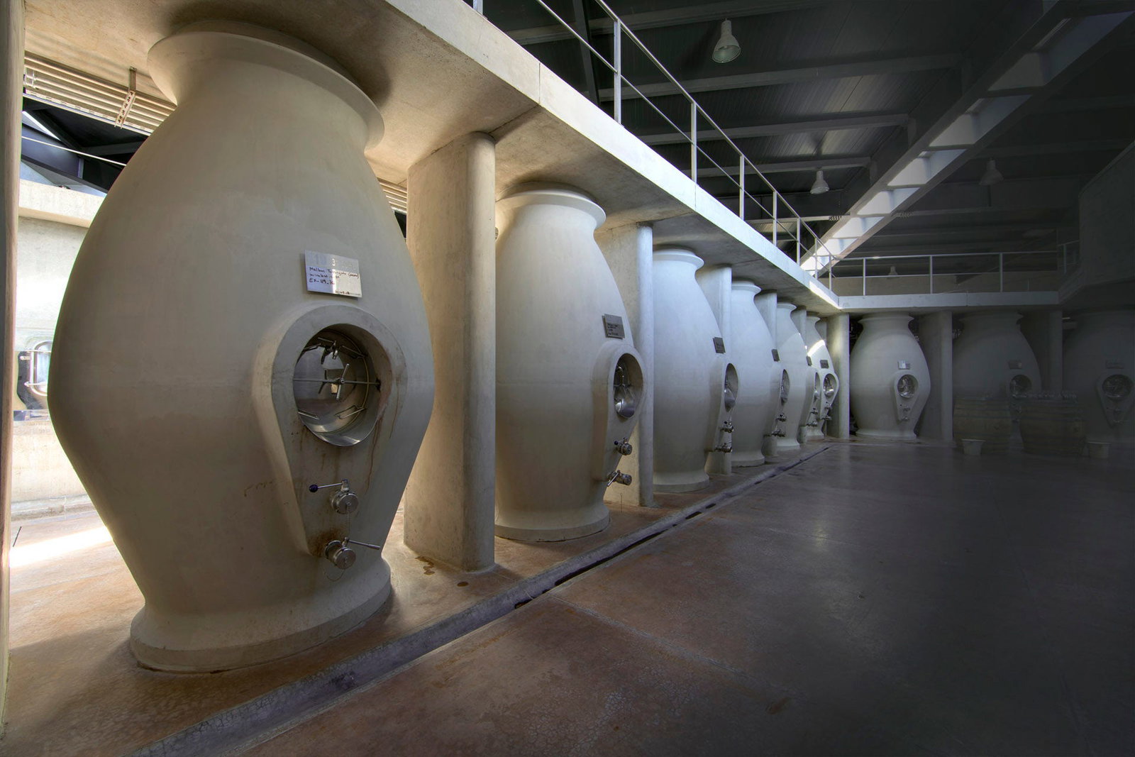 Row of large concrete egg-shaped fermentation/aging vessels in a modern winery cellar with metal walkway above
